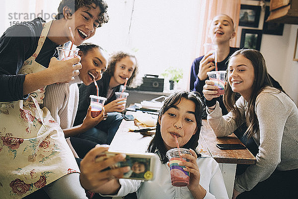 Teenagermädchen nimmt sich mit Freunden über das Mobiltelefon ein  während sie zu Hause einen Smoothie genießt