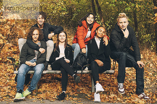 Ganzflächiges Porträt von lächelnden Teenager-Freunden  die im Herbst auf einer Parkbank zusammensitzen