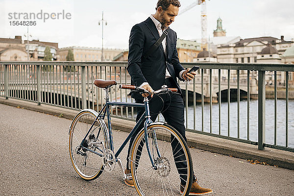 Selbstbewusster Geschäftsmann mit Smartphone beim Gehen mit dem Fahrrad auf der Brücke in der Stadt