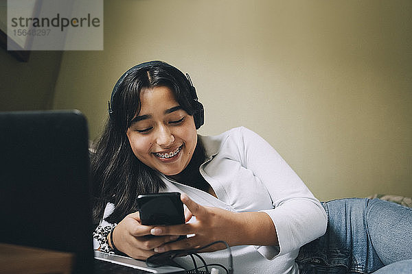 Lächelndes Teenager-Mädchen benutzt Mobiltelefon und Laptop  während es zu Hause im Bett liegt