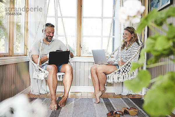 Lächelndes Paar benutzt Laptops  während es auf einer Seilschaukel im Blockhaus sitzt