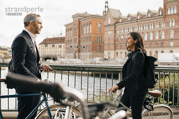 Lächelnde Geschäftsfrau unterhält sich mit Geschäftsmann auf der Brücke  während sie in der Stadt Rad fährt