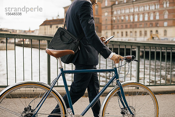 Mittelsektion eines Geschäftsmannes  der ein Smartphone benutzt  während er mit dem Fahrrad auf einer Brücke in der Stadt läuft