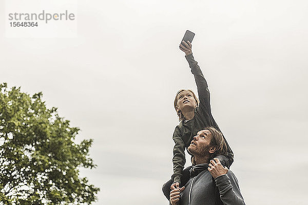 Tochter fotografiert durch ein Smartphone  während sie auf den Schultern des Vaters vor klarem Himmel sitzt