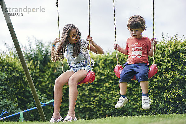 Schwester und jüngerer Bruder auf Schaukeln im Garten