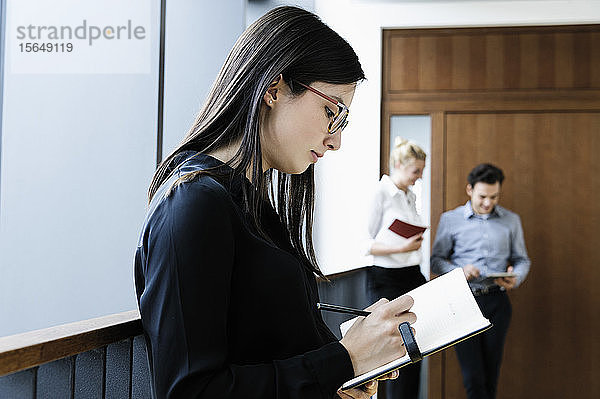 Junge Geschäftsfrau schreibt im Büro Notizen  Kollegen sprechen im Hintergrund