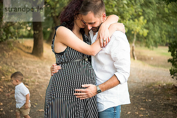 Mann umarmt und berührt den Bauch der schwangeren Frau  Sohn im Hintergrund im Park