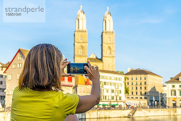 Rückansicht einer Frau  die das Grossmünster und den Fluss Limmat mit einem Smartphone fotografiert  Zürich  Schweiz