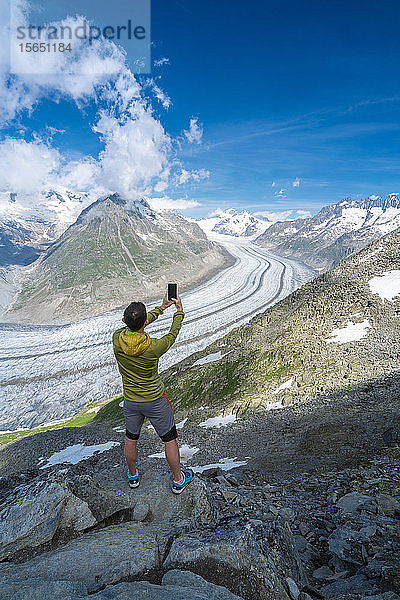 Rückansicht eines Mannes  der den Aletschgletscher mit dem Smartphone vom Aussichtspunkt Eggishorn aus fotografiert  Berner Alpen  Kanton Wallis  Schweiz