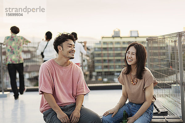 Lächelnde junge Japaner und Japanerinnen sitzen auf einem Dach in einer städtischen Umgebung.