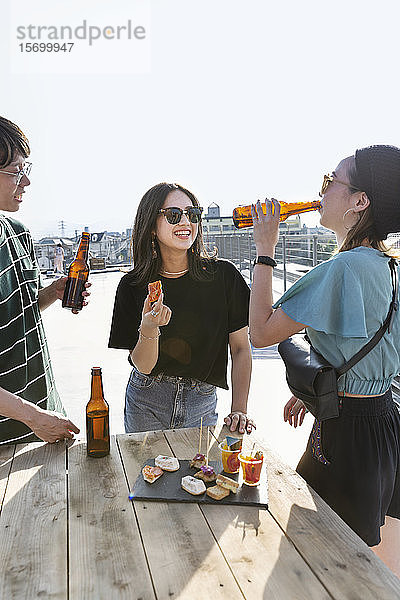 Ein junger Japaner und zwei Frauen stehen auf einem Dach in einer städtischen Umgebung und trinken Bier.