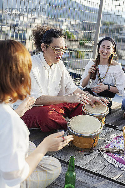 Zwei Japanerinnen und ein Japaner sitzen auf einem Dach in einer städtischen Umgebung und spielen Schlagzeug.
