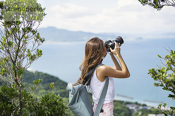Japanische Frau mit Rucksack beim Fotografieren auf einer Klippe  im Hintergrund das Meer.