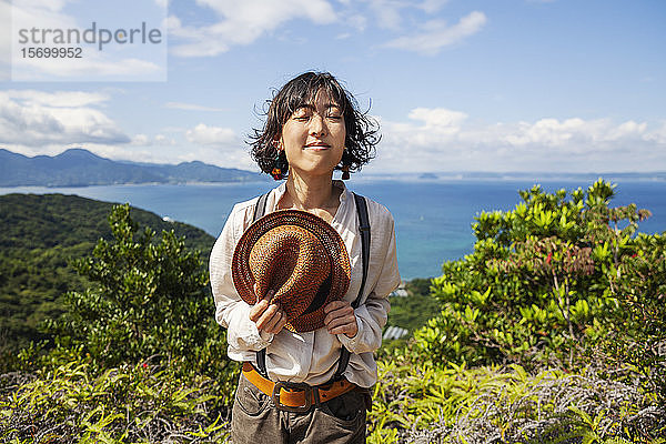 Japanische Frau mit Hut auf einer Klippe stehend  im Hintergrund das Meer.