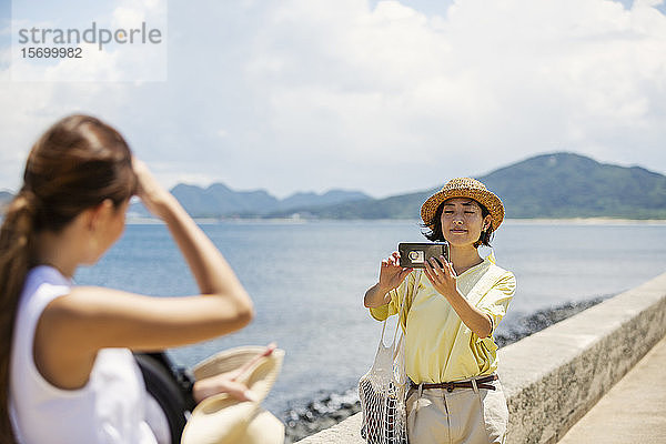 Zwei Japanerinnen stehen am Ozean und fotografieren mit dem Handy.