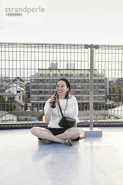 Lächelnde junge Japanerin  die in einer städtischen Umgebung auf einem Dach sitzt und Bier trinkt.