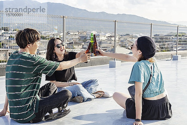 Ein junger Japaner und zwei Frauen sitzen auf einem Dach in einer städtischen Umgebung und trinken Bier.