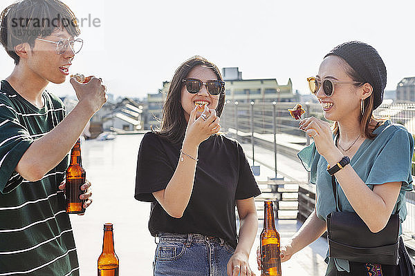 Ein junger Japaner und zwei Frauen stehen auf einem Dach in einer städtischen Umgebung und trinken Bier.
