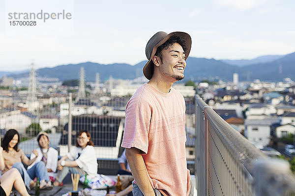 Lächelnder junger Japaner  der auf einem Dach in einer städtischen Umgebung steht.