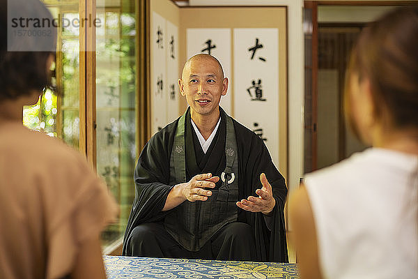 Zwei Japanerinnen und ein buddhistischer Priester knien im buddhistischen Tempel und unterhalten sich.