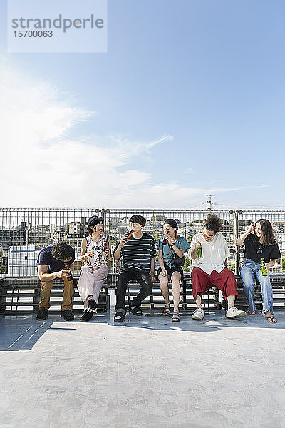 Lächelnde Gruppe junger japanischer Männer und Frauen  die auf einem Dach in einer städtischen Umgebung sitzen.