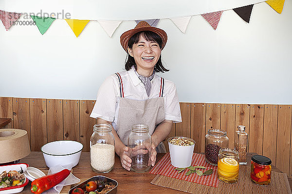 Japanerin mit Hut  die in einem Hofladen mit einer Auswahl an Lebensmitteln und Gewürzen in Gläsern steht.