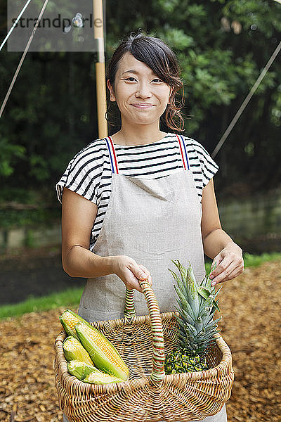 Lächelnde Japanerin mit Schürze steht im Freien  hält Korb mit frischem Gemüse und schaut in die Kamera.