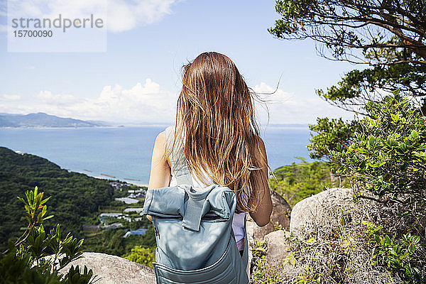 Rückansicht einer Frau mit Rucksack  die auf einem Felsen auf einer Klippe steht  im Hintergrund das Meer.