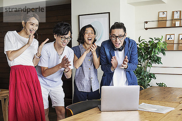 Gruppe junger japanischer Fachkräfte  die sich in einem Raum der Zusammenarbeit lächelnd und jubelnd einen Laptop-Computer anschauen.