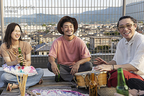 Lächelnde Gruppe junger japanischer Männer und Frauen  die auf einem Dach in einer städtischen Umgebung sitzen.