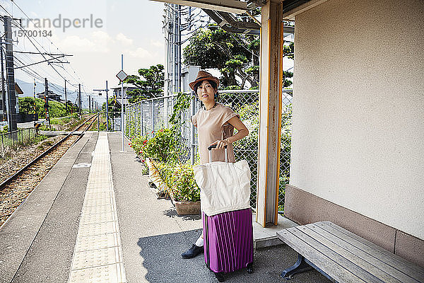 Japanerin mit Hut auf dem Bahnsteig stehend mit Einkaufstasche und rosa Koffer.