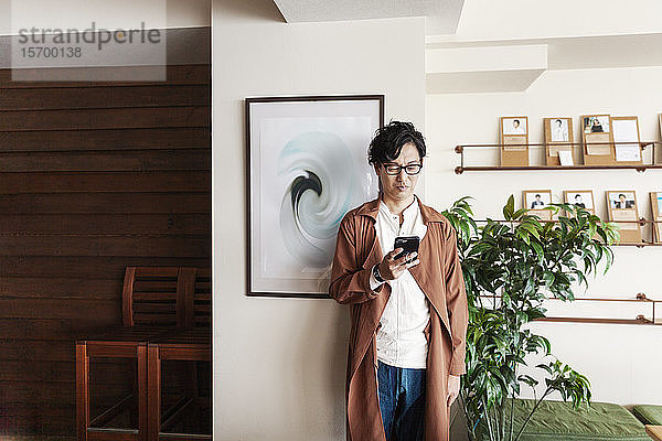 Männlicher japanischer Berufstätiger  der in einem Arbeitsraum steht und auf sein Mobiltelefon schaut.