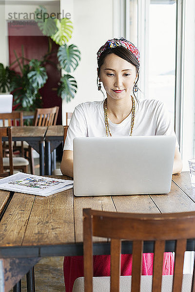 Japanische Fachfrau  die an einem Tisch in einem Arbeitsraum sitzt und einen Laptop benutzt.