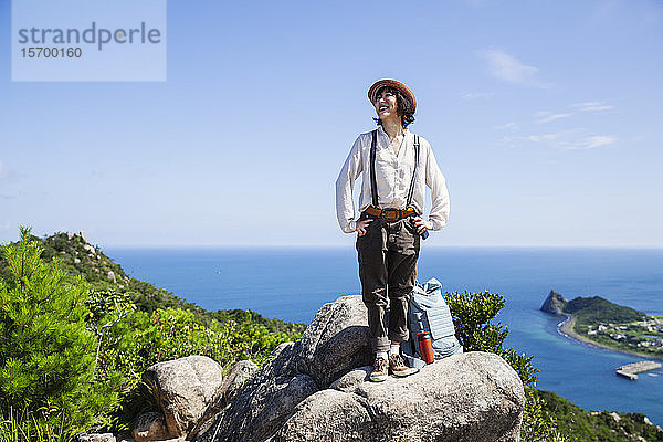 Japanische Frau mit Hut auf einem Felsen auf einer Klippe stehend  im Hintergrund das Meer.