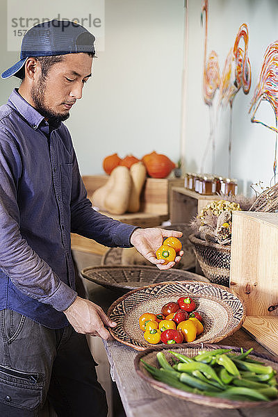 Japanischer Mann mit Mütze steht im Hofladen und hält eine Schüssel mit frischem Gemüse in der Hand.