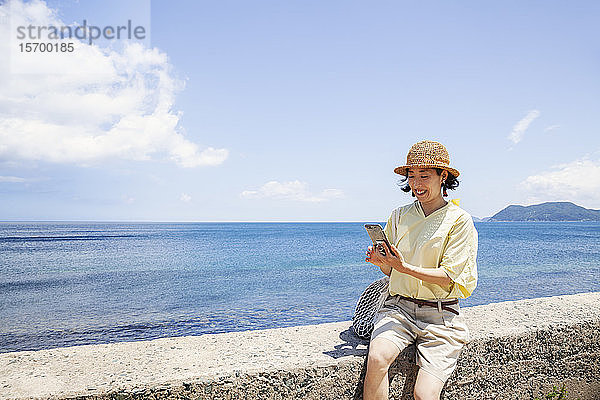 Japanische Frau mit Hut  die an einer Wand sitzt  im Hintergrund das Meer.