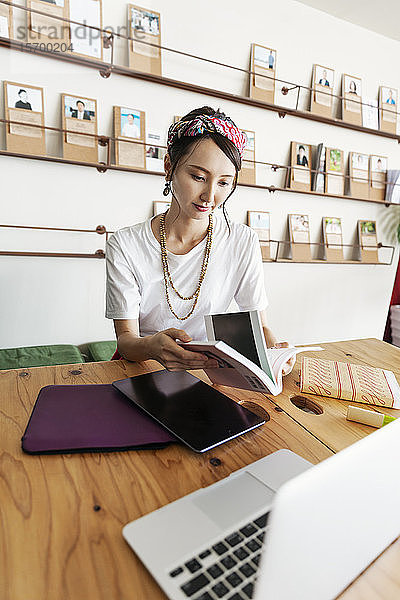 Japanische Fachfrau  die an einem Tisch in einem Raum für Zusammenarbeit sitzt  ein digitales Tablett und einen Laptop benutzt und ein Buch in der Hand hält.