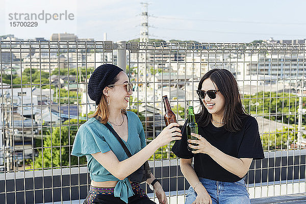 Zwei junge Japanerinnen sitzen auf einem Dach in einer städtischen Umgebung und trinken Bier.