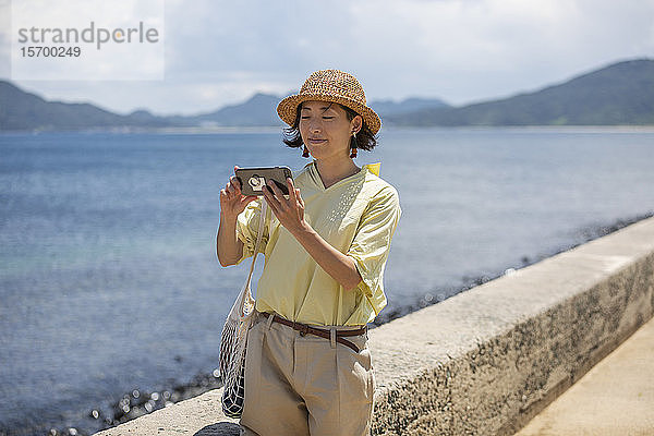 Japanische Frau mit Hut  die am Meer steht und mit dem Handy fotografiert.