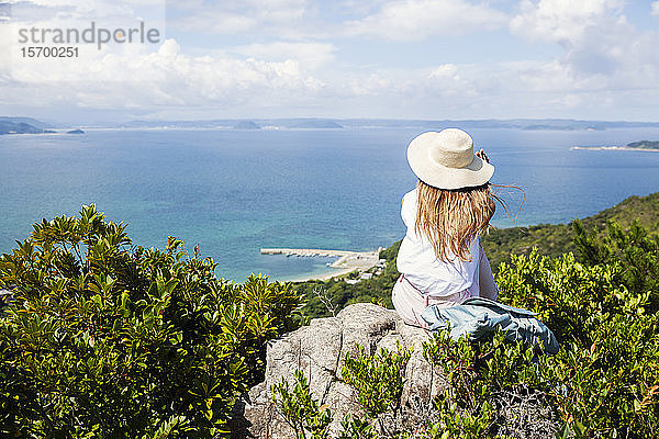 Japanische Frau mit Hut  die auf einem Felsen auf einer Klippe sitzt  im Hintergrund das Meer.