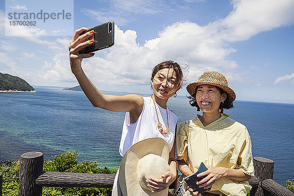 Zwei Japanerinnen mit Hüten auf einer Klippe stehend  Selfie mit Handy  im Hintergrund das Meer.