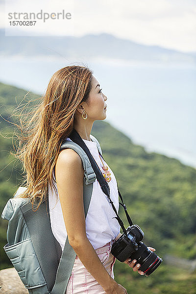 Japanische Frau mit Rucksack und Kamera auf einer Klippe stehend  im Hintergrund das Meer.