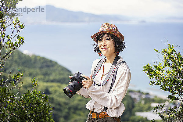 Japanerin mit Hut  Rucksack und Kamera auf einer Klippe stehend  im Hintergrund das Meer.