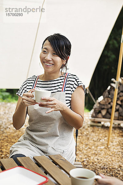Lächelnde Japanerin  die draußen an einem Tisch sitzt und eine Tasse Kaffee trinkt.