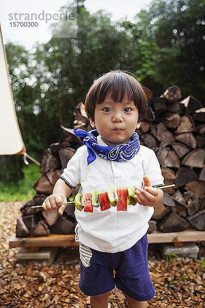 Porträt eines japanischen Jungen  der einen Spieß mit frischer Kiwi und Melone hält und in die Kamera schaut.
