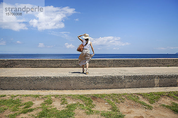 Japanische Frau mit Hut an der Wand stehend  im Hintergrund das Meer.