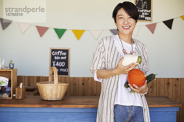 Japanische Frau steht in einem Hofladen  hält Gemüse und lächelt in die Kamera.