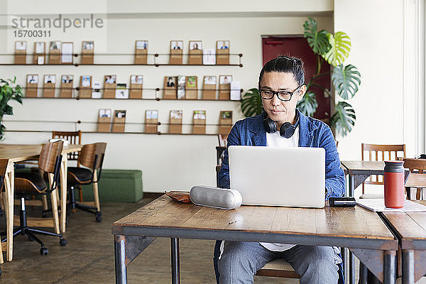 Männlicher japanischer Berufstätiger  der an einem Tisch in einem Raum für Zusammenarbeit sitzt und einen Laptop-Computer benutzt.