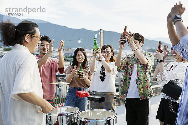 Lächelnde Gruppe junger japanischer Männer und Frauen  die auf einem Dach in einer städtischen Umgebung stehen und Bier trinken.