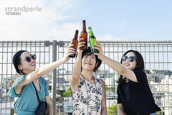 Drei junge Japanerinnen sitzen auf einem Dach in einer städtischen Umgebung und trinken Bier.